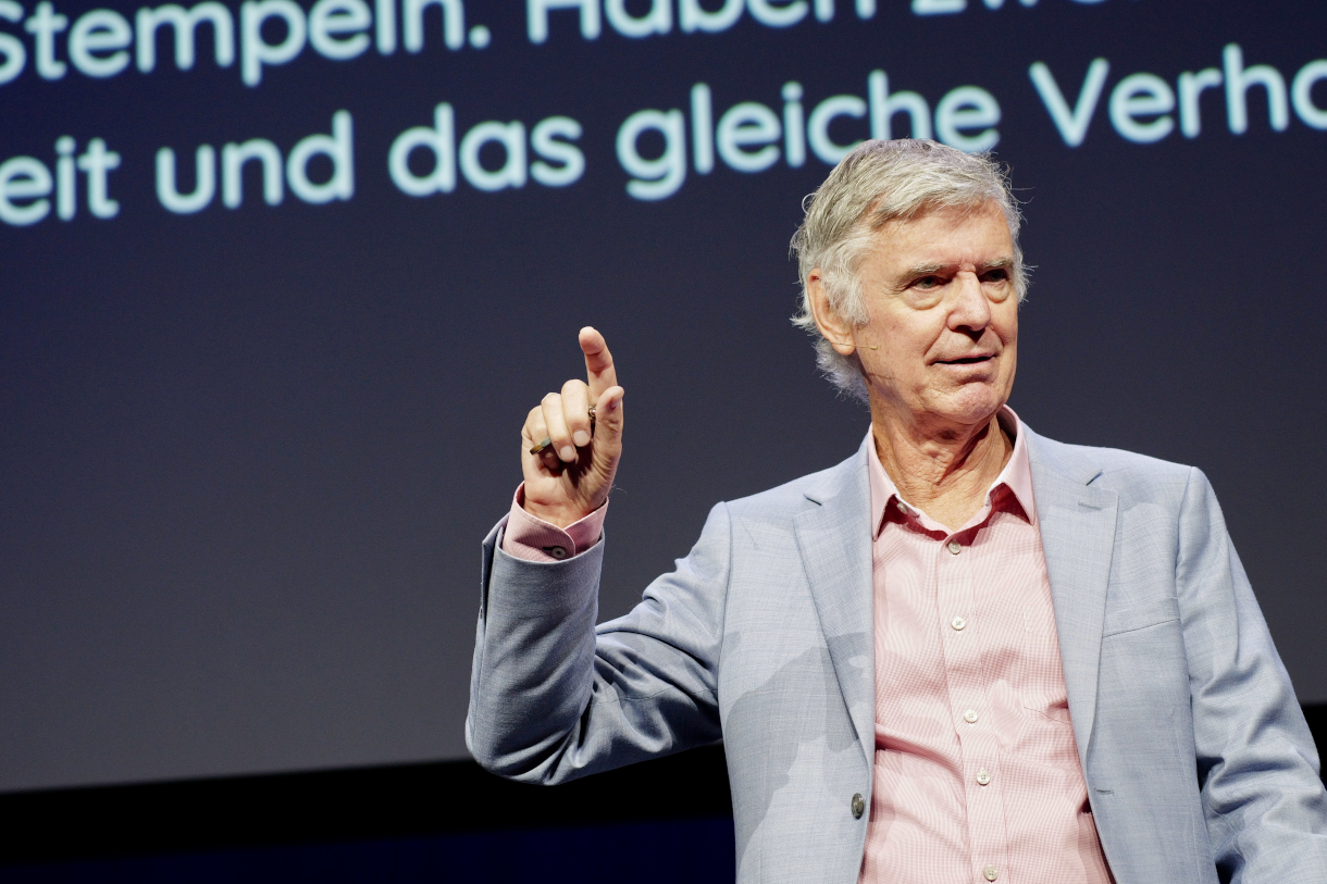 Foto: Professor Dr. h. c. John Hattie steht bei seinem Vortrag auf dem ersten Schulleitungssym-posium Baden-Württemberg in Heilbronn vor einer Powerpoint-Präsentation, von der nur wenige Buchstaben zu erkennen sind (lesbar sind nur „Stempeln … und das gleiche Verhalten“). Hattie hebt gerade einen Arm und deutet mit seinem Zeige-finger vor sich in die Luft. Hattie hat graue Haare und trägt ein graublaues Jacket und ein roséfarbenes Hemd.