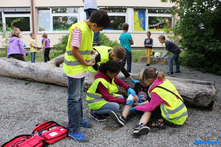 Symbolbild für Juniorhelferprogramm (Foto: vier Kinder in Warnwesten leisten Erste Hilfe bei einem Jungen auf dem Pausenhof einer Grundschule; im Hintergrund spielen acht Kinder mit einem Ball)