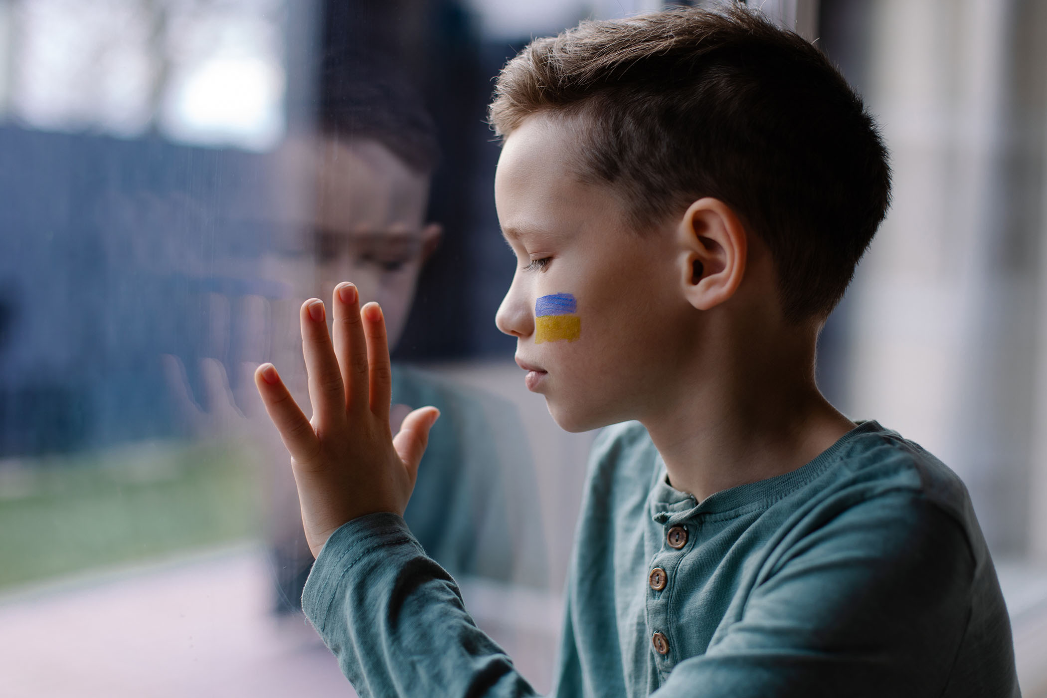 Foto: Junge, der eine ukrainische Flagge auf die Wange gemalt hat, blickt traurig aus dem Fenster.