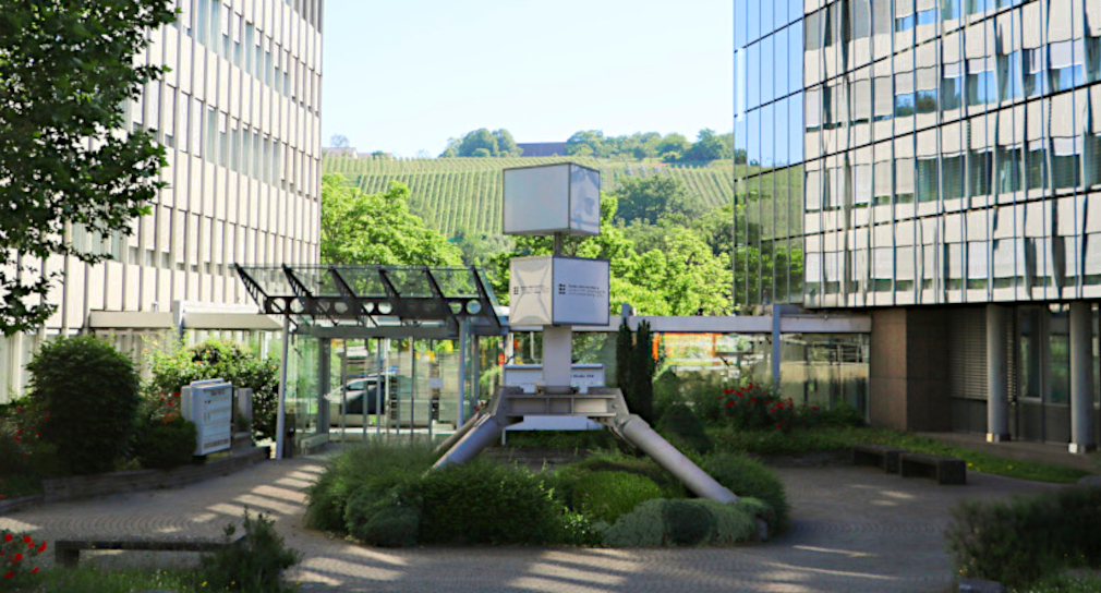 Foto: Gebäude der ZSL-Zentrale in Stuttgart: Im Vordergrund in der Mittel steht auf einem kleinen Vorplatz eine Säule mit zwei Würfeln; auf dem unteren Würfel ist die Wort-Bild-Marke des ZSL zu sehen. Links und rechts sieht man die beiden mehrstö-ckigen Hauptgebäude, die in der Mitte durch einen Flachbaugang miteinander verbun-den sind. Im Hintergrund sieht man Weinberge.