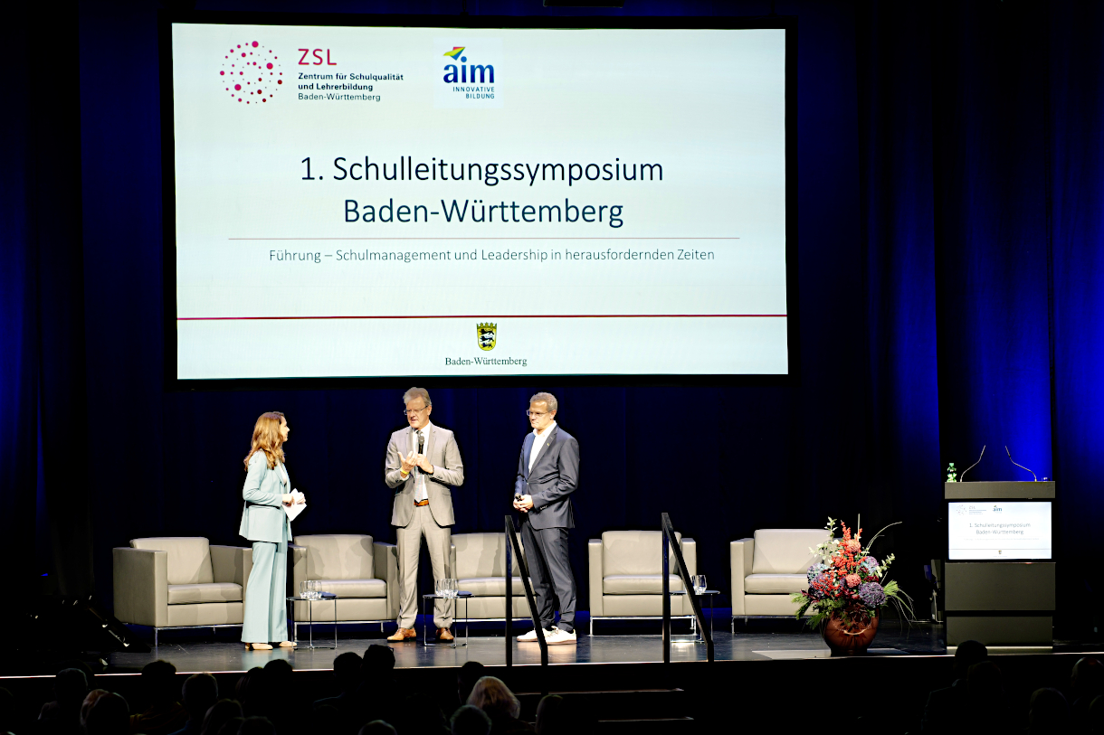 Foto: Begrüßung zum 1. Schulleitungssymposium Baden-Württemberg (auf dem Podium von links nach rechts stehen die Moderatorin, ZSL-Präsident Prof. Dr. Thomas Riecke-Baulecke und aim-Geschäftsführer Marco Haaf, im Hintergrund ist vor ei-nem dunkelblauen Vorhang eine Präsentationsfolie mit dem Titel der Veranstaltung und den Logos von ZSL und aim zu sehen; vor dem Podium sieht man einige Köpfe von Zuhörenden)