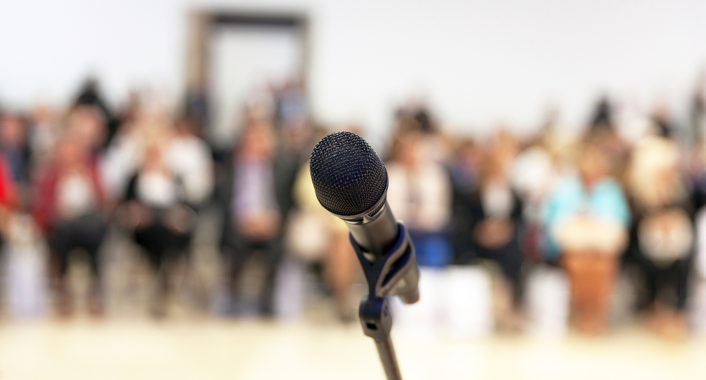 Symbolbild für Schulpreis: Mikrofon vor unscharfem Hintergrund mit Publikum