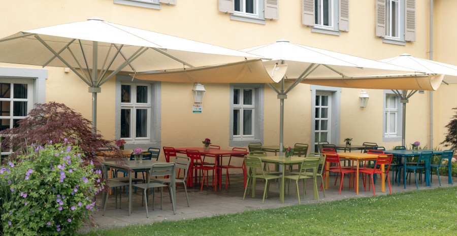 Foto: Blick vom Rasen auf eine Terrasse vor einer historischen Fassade, mit farbigen Tischen und Stühlen, überdacht von cremeweißen großen Schirmen.