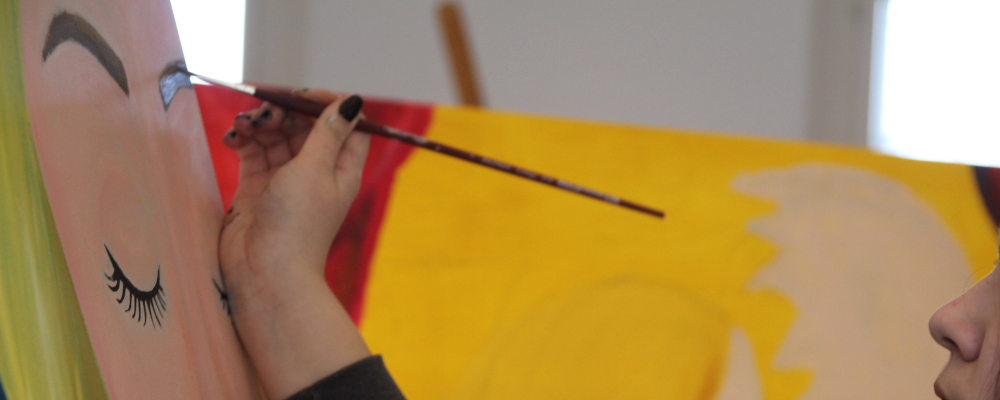 Foto: Ausschnitt aus einer Ateliersituation, in der ein junges Mädchen an einer Staffelei steht und ein Frauengesicht malt.