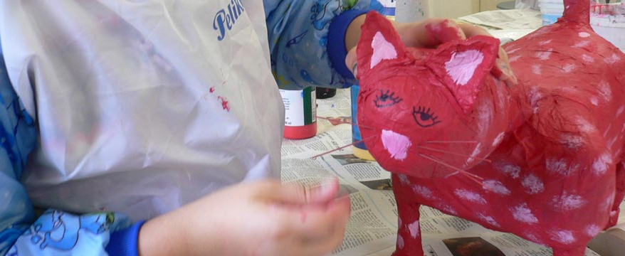 Foto: Ein Kind im Malkittel bearbeitet eine rote Pappmaché-Katze mit rosa Punkten.