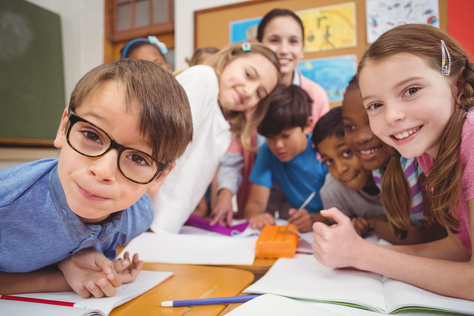Symbolbild für Schulpsychologie (Foto: circa acht Grundschülerinnen und Grundschüler stehen um einen Tisch herum im Klassenzimmer; im Hintergrund steht die Lehrerin; alle schauen lächelnd in die Kamera)