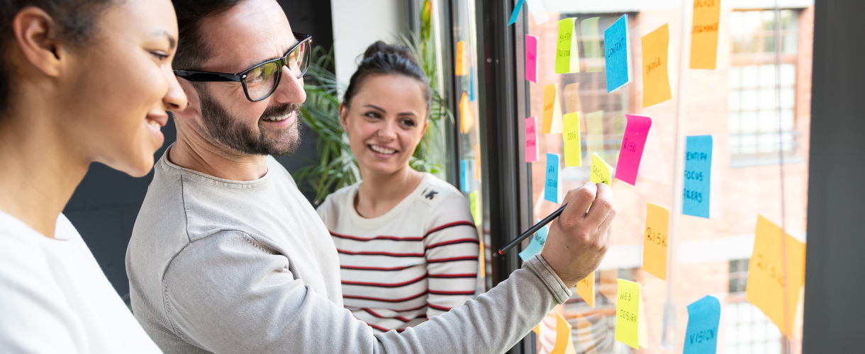 Link zur ZSL-Aktuelles-Meldung: Informationsveranstaltung Weiterbildungsmaster Schulmanagement und Leadership (5. Mai und  9. Juli 2026, jeweils 18:30 bis 19:30 Uhr) (Foto: Zwei Frauen und ein Mann stehen vor einem Fenster, auf dem farbige Post-its kleben. Der Mann schreibt mit einem Stift etwas auf einen der Zettel. Alle drei Personen lächeln.)