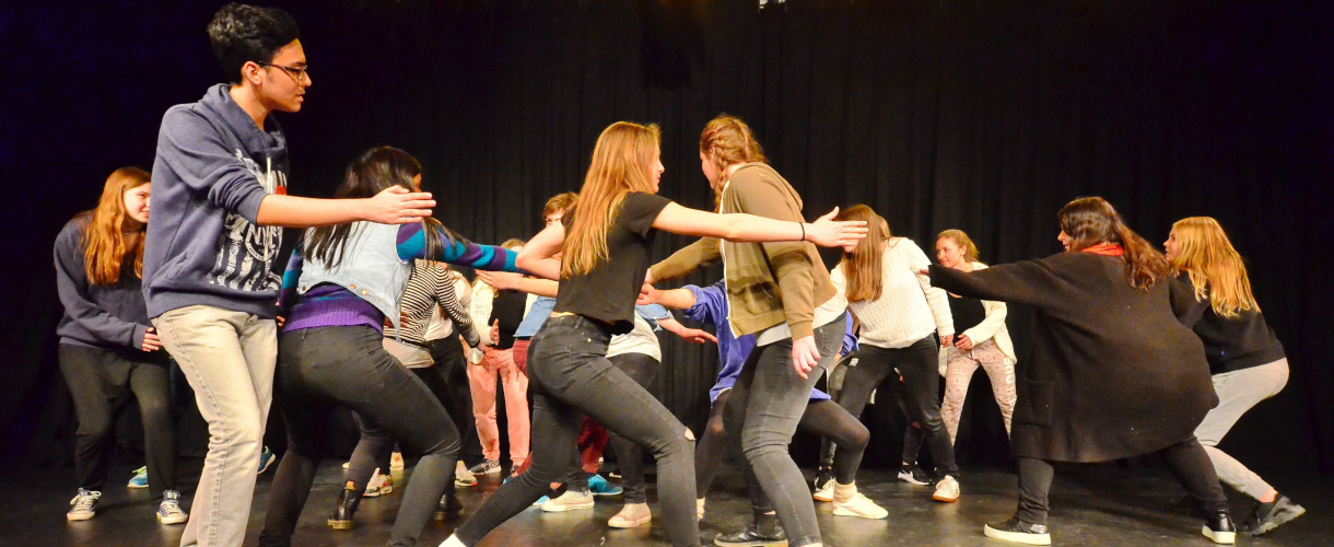 Foto aus einem Theaterworkshop: Eine Gruppe Jugendlicher bewegt sich mit großen Schritten und schwingenden Armen durch einen Raum.