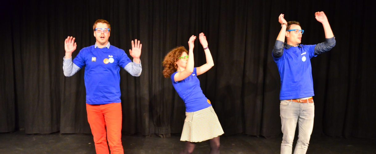 Foto einer Szene: Auf einer Bühne mit schwarzem Hintergrund stehen drei Personen in blauen T-Shirts und strecken ihre Arme in die Höhe.