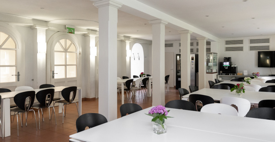 Foto: Blick durch einen länglich angelegten Raum, der mit Holzpfeilern gegliedert und mit großen weißen Gruppentischen eingerichtet ist.