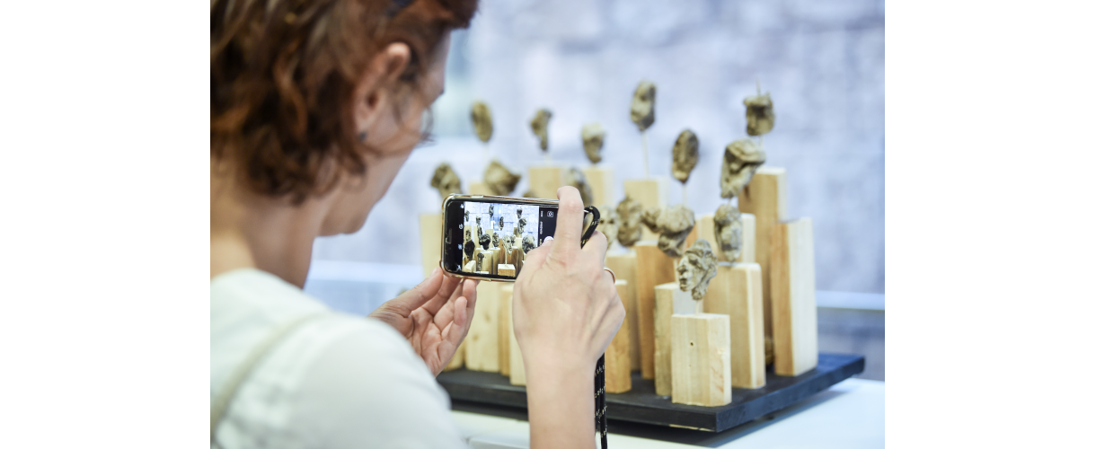 SCHULKUNST-Landesausstellung „Serie“ (Foto: Frau, deren Kopf und Oberkörper von hinten zu sehen ist, fotografiert mit ihrem Smartphone ein Kunstobjekt. Dieses besteht aus mehreren senkrechte stehenden Holzrechtecken, auf die kleine Kopfskulpturen mit Draht aufgesteckt sind.)