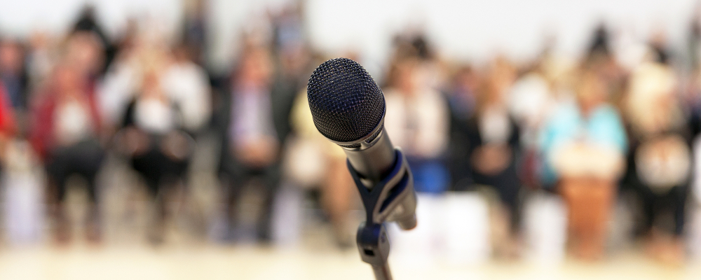 Symbolbild zu Schulpreis „Demokratie und Werte (er)leben!“ (Foto: Mikrofon vor unscharf gestelltem Hintergrund sitzender Menschen)