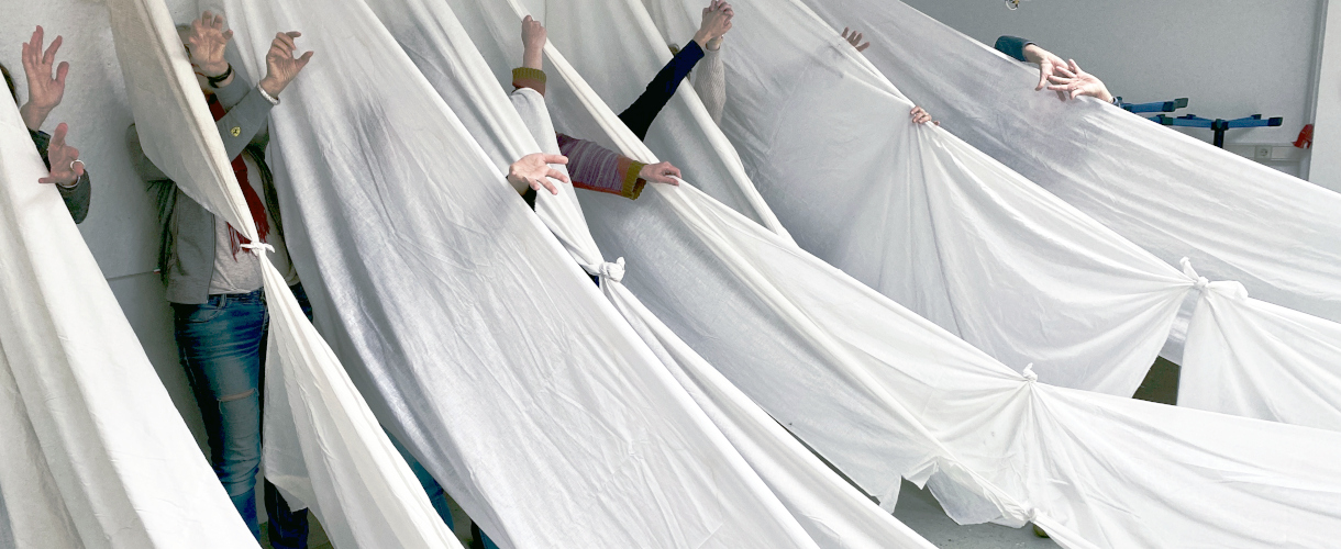 Foto: Mehrere Personen bewegen sich hinter aufgehängten weißen, langen Stoffbahnen, aus denen nur ihre Arme und Beine hervorragen – Teil einer performativen Gruppenarbeit im Raum.