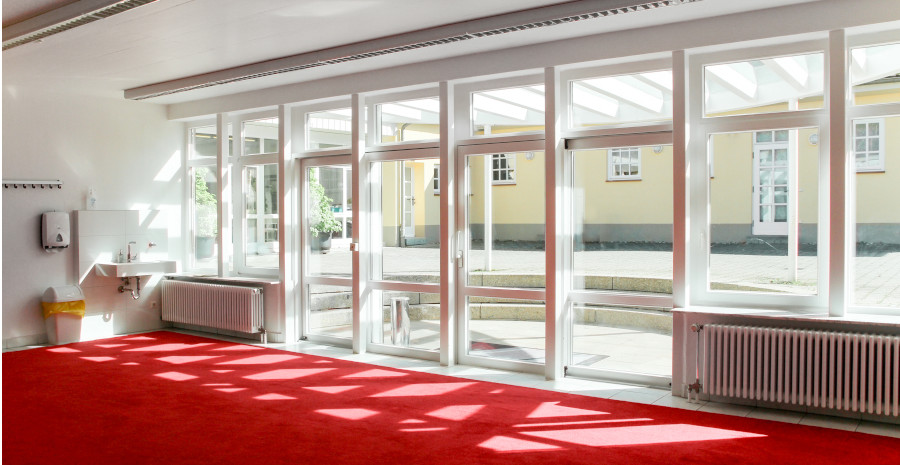 Foto: Blick aus einem Raum mit rotem Teppich durch eine verglaste Wand in ein glasüberdachtes Atrium.