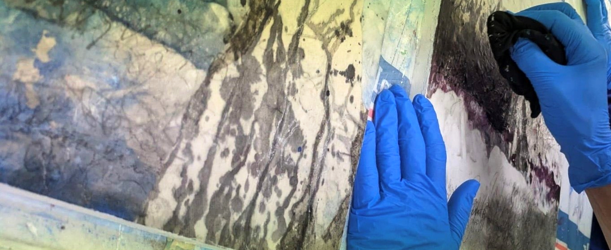 Foto: Zwei Hände mit blauen Gummihandschuhen bearbeiten mit einem schwarzen Tuch eine abstrakte, marmorierte Oberfläche mit dunklen und hellen Farbbereichen.