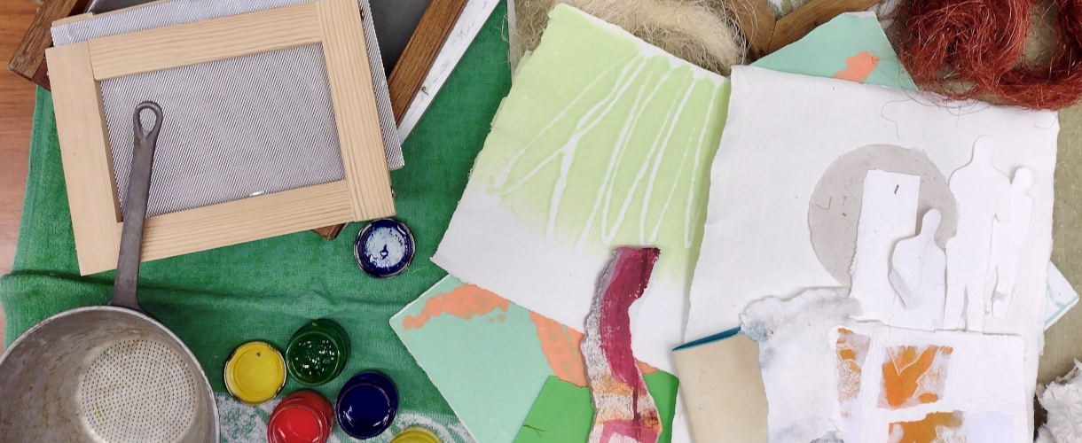 Foto: Verschiedene Materialien zum künstlerischen Arbeiten liegen auf einer Stoffunterlage, darunter Holzrahmen mit Siebgewebe, Farbtöpfchen, Papier und Naturfasern, von oben fotografiert.