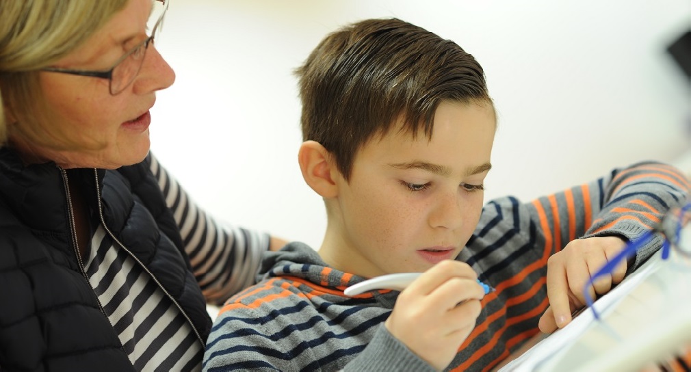 Symbolbild Unterstützungs- und Förderbedarfe: Ein Junge schreibt konzentriert in sein Heft und die Lehrerin schaut ihm dabei aufmerksam über die Schulter.