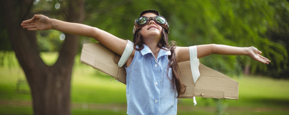 Symbolbild für Startchancen-Programm BW: Ein Mädchen steht auf einer grünen Wiese. Im Hintergrund ist ein Baum zu sehen. Das Mädchen trägt eine Fliegerbrille, Pappflügel, ein hellblaues mit Kragen und dunkelblaue Jeans. Es schaut nach oben und hat die Arme so ausgestreckt, als wolle es fliegen.