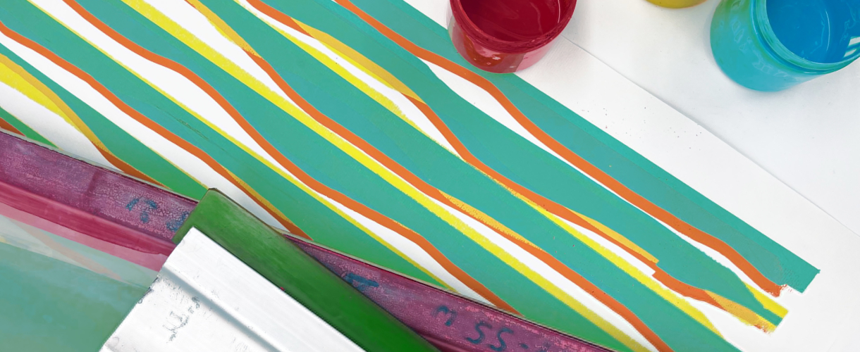 Foto: Nahaufnahme einer Siebdruckgrafik mit geschwungenen, parallelen Streifen in den Farben Grün, Orange und Gelb. Im Vordergrund links ist ein Ausschnitt eines Siebdruckrahmens mit Rakel zu sehen, rechts oben stehen Farbtöpfe in den Farben Rot und Blau.