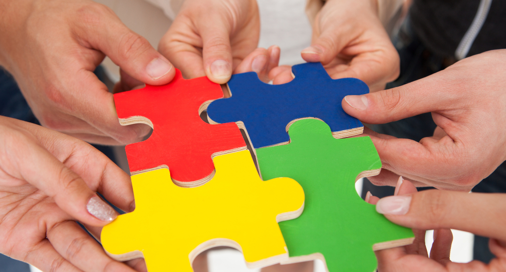 Symbolbild Schulpsychologische Dienste: Sechs Hände halten vier große Puzzle-Teile. Die Puzzle-Teile tragen unterschiedliche Farben, nämlich rot, blau, gelb und grün. Sie passen einwandfrei ineinander. 