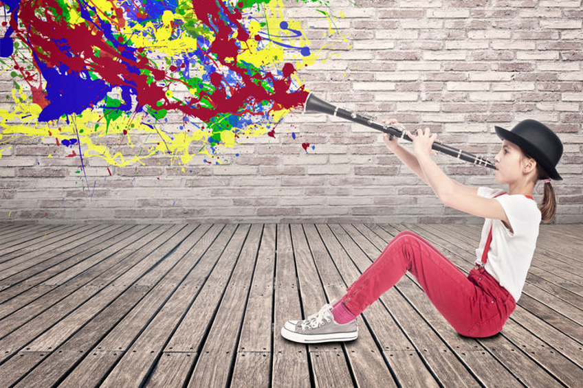 Musik (Foto mit grafischen Elementen: Ein auf dem Holzboden sitzendes Mädchen mit Hut, in roter Hose und weißem T-Shirt, spielt Klarinette. Sie hält das Instrument schräg in die Höhe. Aus dem Schallloch strömt eine Wolke bunter Farben.)