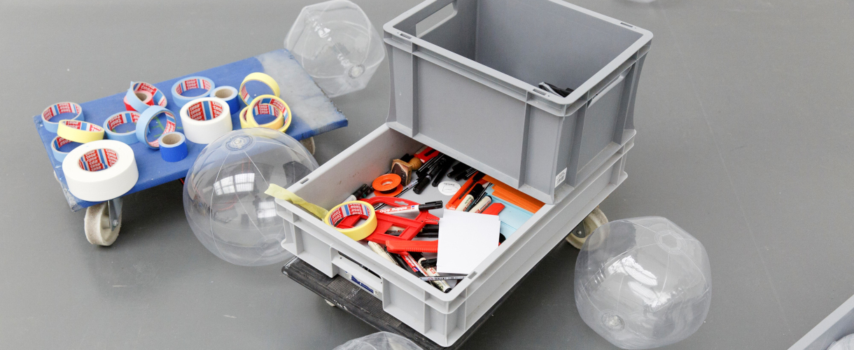 Foto: Materialien für den Kunstunterricht wie Klebeband, Stifte, Scheren, Cutter und transparente aufblasbare Kunststoffkugeln liegen sortiert auf einem Rollbrett und in einer grauen Kiste auf dem Boden.
