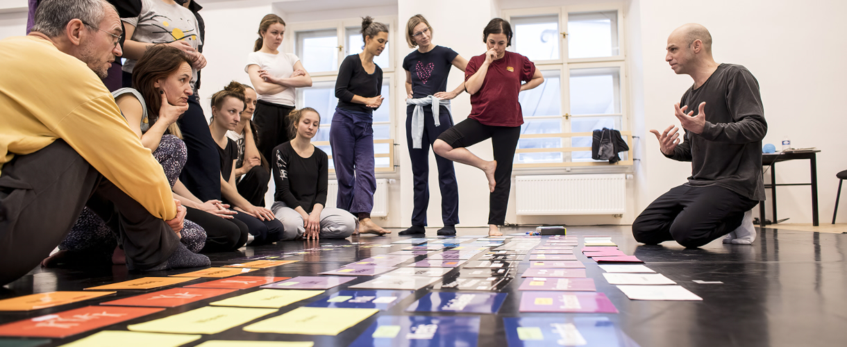 Foto aus einem Theaterworkshop: die Workshopleitung kniet auf dem Boden und erklärt verschiedene, auf dem Boden liegende Karten, die für die Entwicklung von Tanzchoreografien verwendet werden, während die Teilnehmenden zuhören.