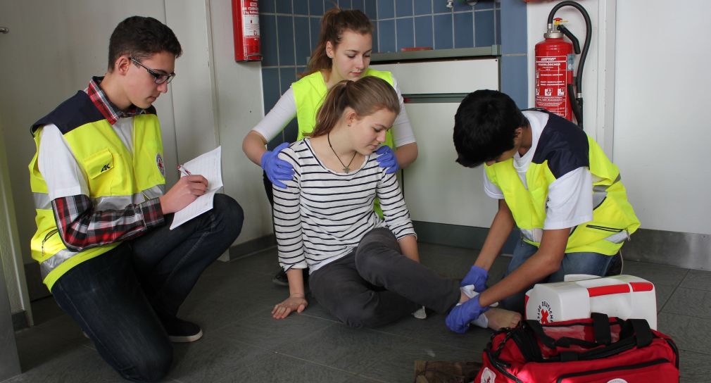 Link; Symbolbild Erste-Hilfe-Maßnahmen für Schülerinnen und Schüler (Foto: Drei Schüler in gelben Warnwesten mit einem geöffneten Notfallkoffer kümmern sich um eine verletzte Schülerin.)