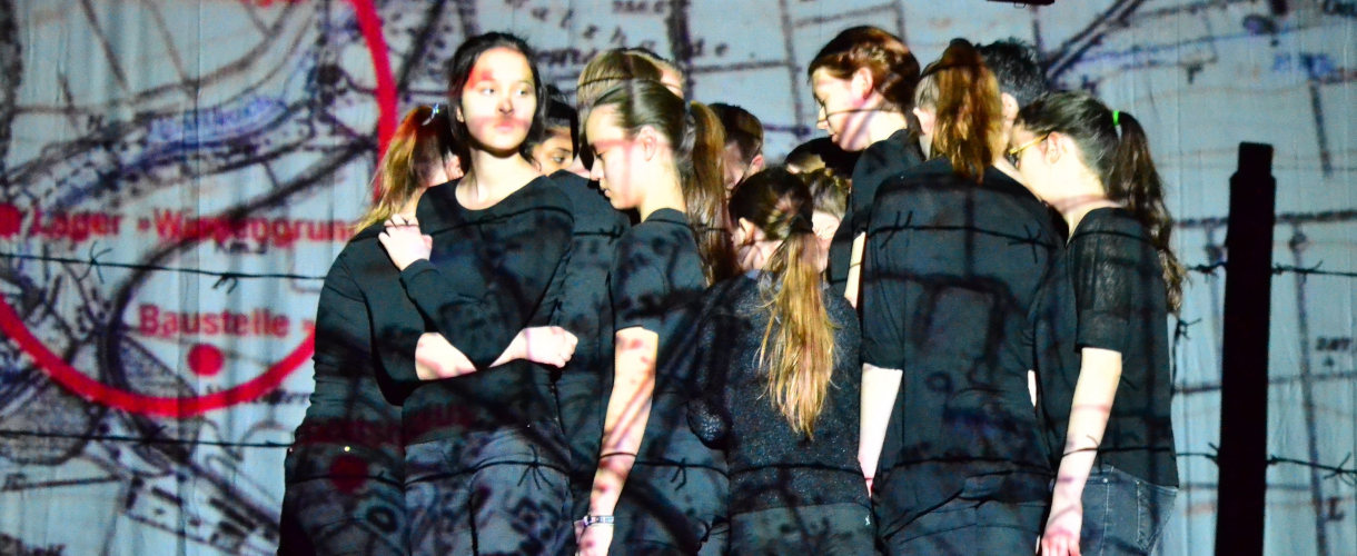 Foto einer Szene: Vor einer videoprojizierten Landkarte bildet eine Gruppe Menschen in schwarzer Kleidung einen Pulk.