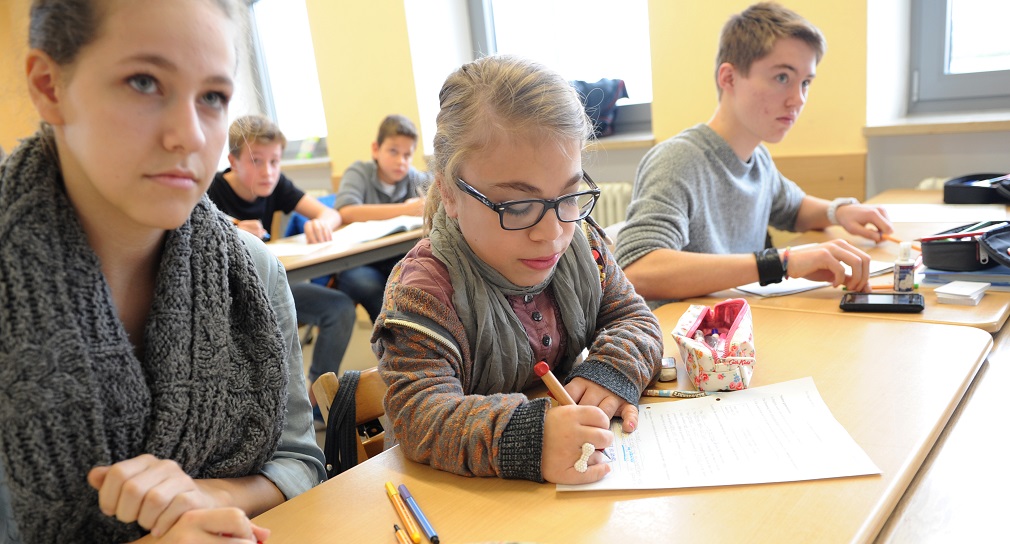 Link; Symbolbild Kooperation allgemeine Pädagogik und Sonderpädagogik (Foto: Schüler sitzen im Klassenraum; ein Mädchen mit Brille schreibt konzentriert, während andere aufmerksam mitarbeiten.)