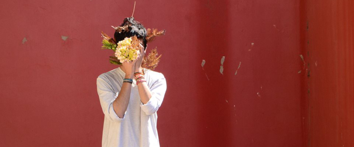 Foto einer Szene: Vor einem roten Hintergrund steht eine Person mit schwarzen Haaren, in einem weißen Hemd und hält sich eine Strauß Blüten vor das Gesicht.