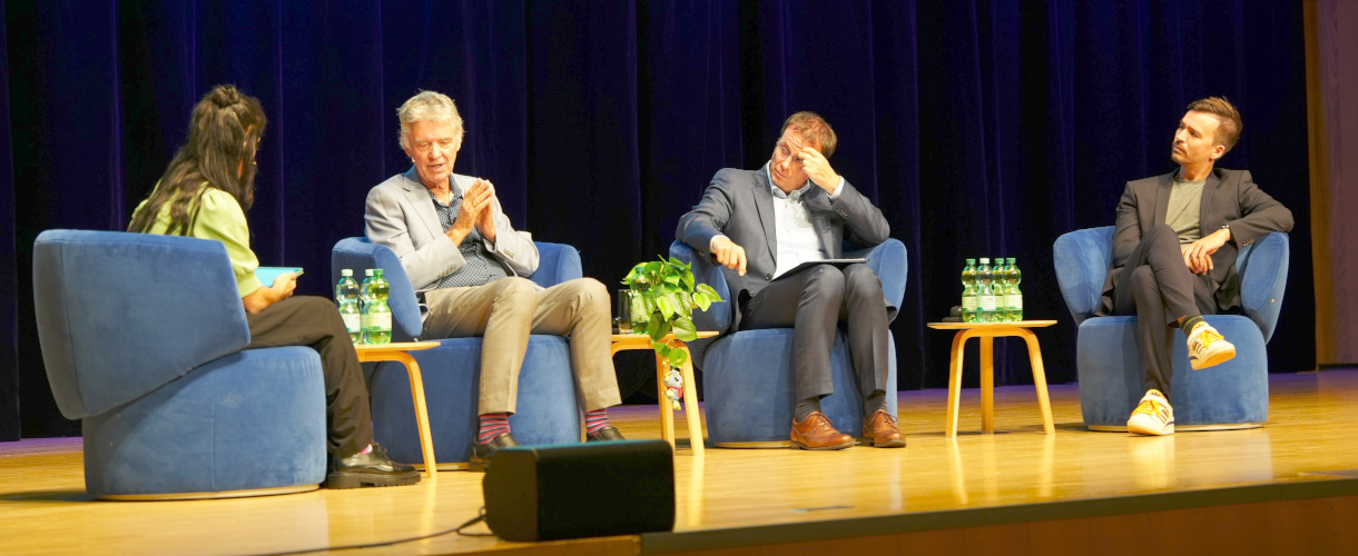 Prof. John Hattie im Gespräch mit der Moderatorin, Staatssekretär Volker Schebesta und Bildungs-Influencer Bob Blume bei der Abendveranstaltung „Mit Visible Learning zum Lernerfolg“ (Heilbronn, 22.09.2025) (Foto: Die Diskussionsteilnehmenden sitzen auf blauen Sesseln auf der Bühne. Von links nach rechts sind zu sehen: Moderatorin, John Hattie, Volker Schebesta und Bob Blume. Hattie spricht mit der Moderatorin.)