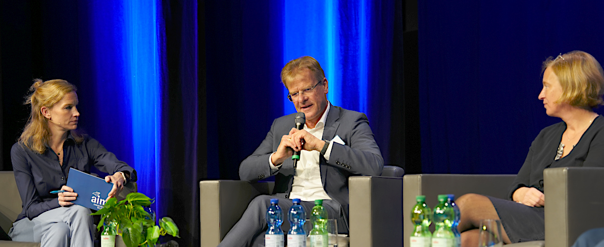 Prof. Dr. Thomas Riecke-Baulecke spricht beim Panel der Fachtagung: „Lernen sichtbar machen – Chancen pädagogischen Handelns in Kita und Grundschule“ am 23.09.2025 in Heilbronn (Foto: von links nach rechts: Moderatorin, Prof. Dr. Riecke-Baulecke, Prof. Dr. Christina Schwer; nicht im Bild: Prof. John Hattie, der rechts neben Prof. Dr. Schwer sitzt. Alle sitzen auf der Bühne in eckigen Sesseln. Prof. Dr. Riecke-Baulecke spricht in ein Mikro.)