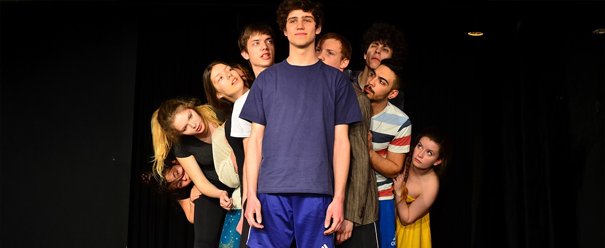 Foto einer Szene: Eine Person steht vor schwarzem Hintergrund lächelnd in der Mitte und hinter ihr stehen viele Personen, die sie neugierig und erstaunt anschauen.