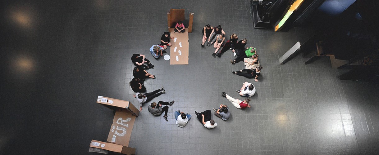 Foto einer Theatergruppe: Eine Jugendliche sitzt auf einem Pappkarton voller weißer Fußabdrücke, um den sich ein Sitzkreis mit weiteren Jugendlichen bildet.