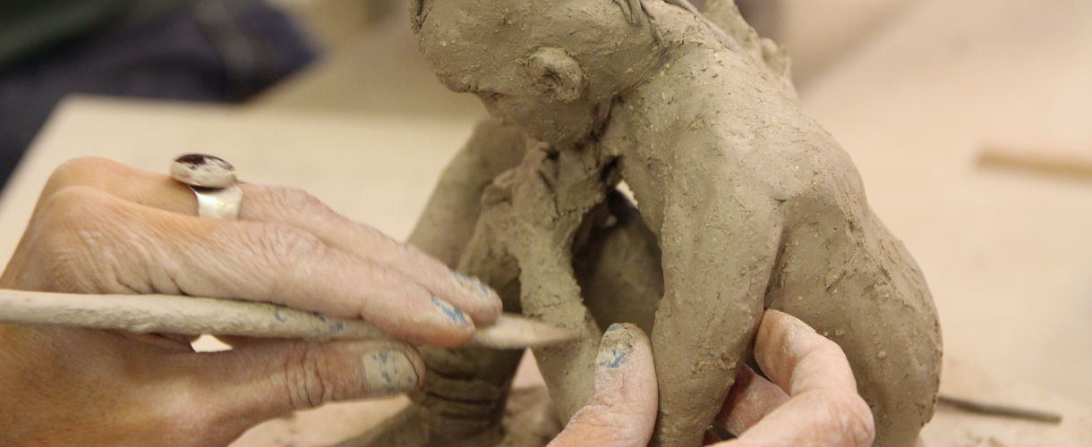 Foto: Eine Hand formt mit einem Modellierwerkzeug Details an einer sitzenden menschlichen Tonfigur. 