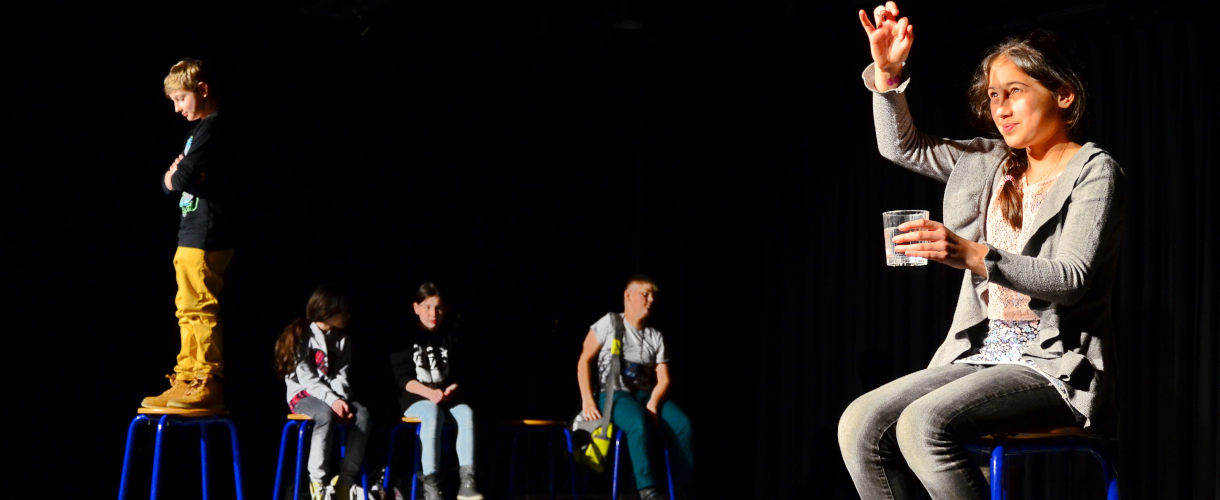 Foto einer Szene: Vor einem schwarzen Hintergrund stehen und sitzen 5 Kinder auf Hockern und das Mädchen im Vordergrund hält ein Glas mit Wasser in der linken Hand und betrachtet ihre rechte Hand, die sie auf Höhe ihres Gesichts hält.