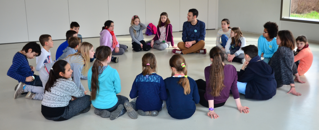 Foto aus einem Theaterworkshop: Eine Gruppe Kinder sitzt, mit einem Theaterpädagogen ins Gespräch vertieft, in einem Kreis.