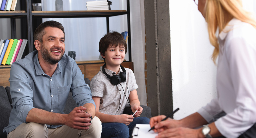 Symbolbild Beratungslehrkräfte: Ein Vater und ein Sohn hören einer Beratungslehrkraft aufmerksam zu. Die Beratungslehrkraft macht sich Notizen auf einem Klemmbrett.