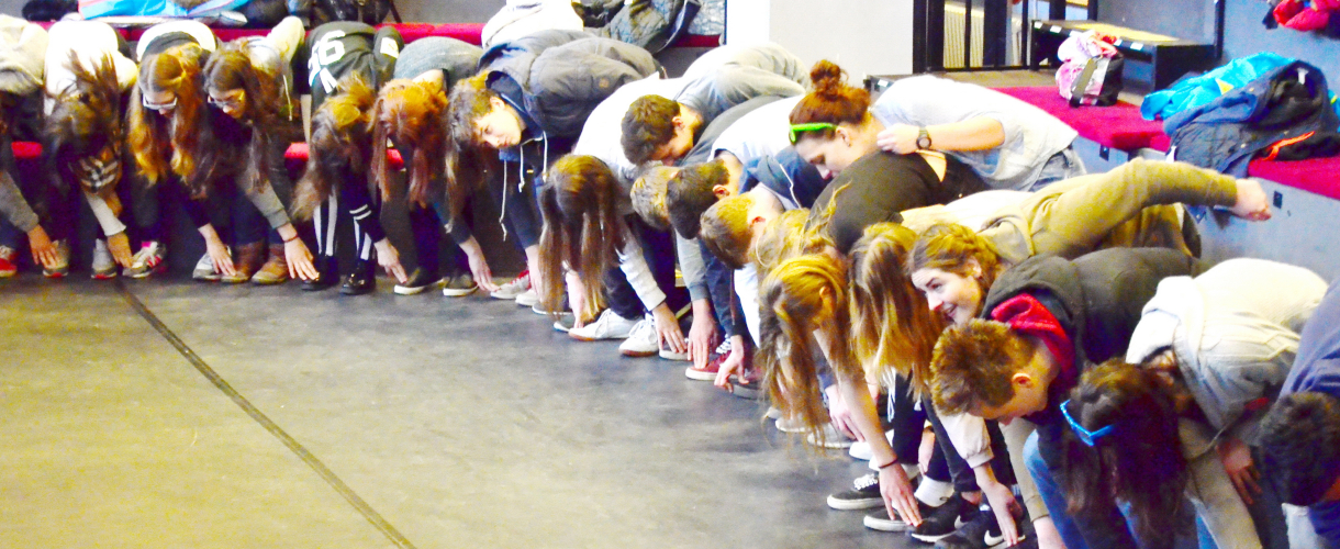 Foto aus einem Theaterworkshop: Eine Reihe Menschen beugt sich vornüber und berührt mit der rechten Hand den linken Fuß der danebenstehenden Person.