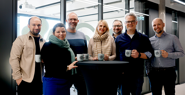 Foto: Auf dem Foto ist das Kernteam des KI-Zentrums Schule zu sehen: Folgende Personen stehen halbkreisförmig um einen Stehtisch herum und halten Tassen mit IPAI-Logo, von links nach rechts: Patrick Hegedüs, Irmi Mühlhuber, Michael Brenner, Annely Humpreys, Sven Kaufmann, Benedikt Sauerborn, Michael Dohmen.