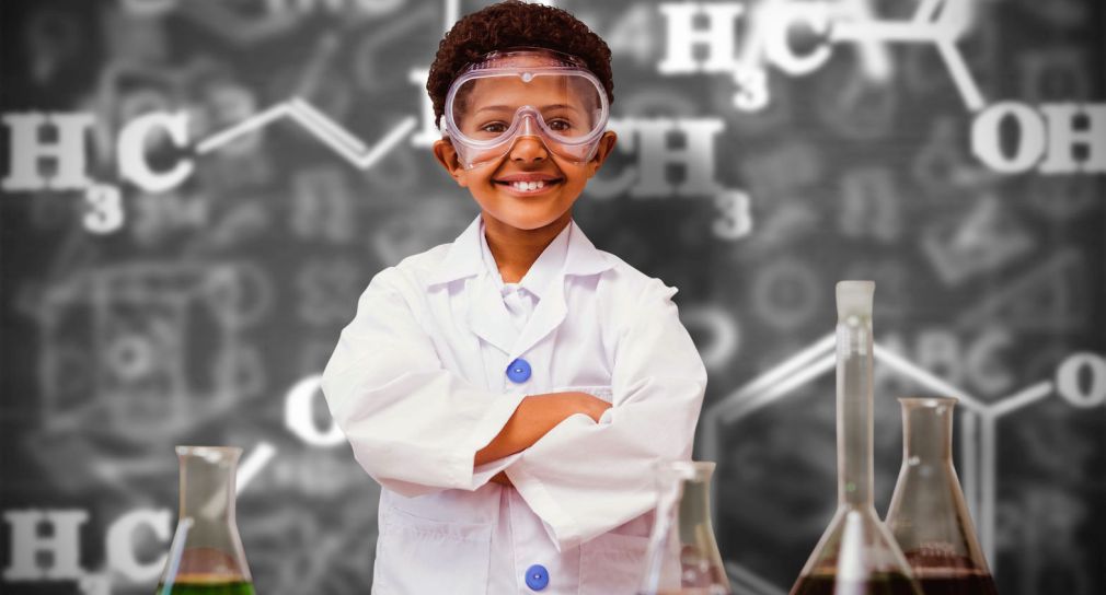 Symbolbild für Zertifikatsstudium: Foto: Junge steht in Chemielabor (Der kleine Junge trägt einen weißen Laborkittel und eine für sein Gesicht sehr große Laborbrille. Er hat die Arme verschränkt und lächelt die Bildbetrachtenden an. Im Hintergrund ist eine Tafel mit chemischen Formeln zu sehen. Neben dem Jungen stehen links und rechts Laborglasgefäße.)