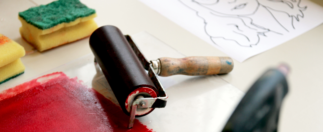 Foto: Materialien für den Kunstunterricht, darunter eine Farbwalze mit roter Farbe, Schwämme und eine gezeichnete Porträtvorlage auf Papier.