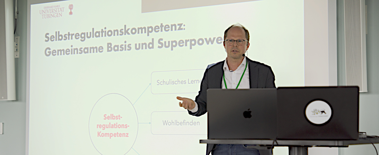 Foto vom 29. Juli 2025, Fachtag „Selbstregulation – zentrale Voraussetzung für erfolgreiches Lernen und Lehren“ am KI Zentrum Schule in Heilbronn: Prof. Dr. Ulrich Trautwein hält einen Vortrag. Er steht an einem Pult mit zwei Laptops vor einer PowerPoint-Präsentation mit dem Folientitel „Selbstregulationskompetenz: Gemeinsame Basis und Superpower“.