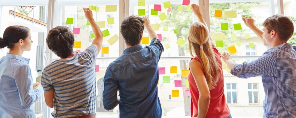 Symbolbild: Kriterien sammeln (Foto: Fünf Personen, die dem Betrachter den Rücken zuwenden, kleben Post-its an eine Fensterfront.)