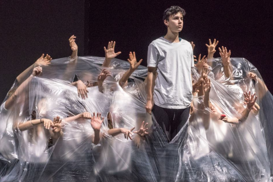 Link zur ZSL-Seite „Theater und Schultheater“; Theaterszene (Foto: Vor schwarzem Hintergrund ist eine große durchsichtige Folie zu sehen, unter der Menschen knien oder hocken. Man kann ihre Gesichter nicht erkennen. Sie strecken ihre Arme und Hände durch Risse in der Folie nach außen. Zentral sichtbar steht eine junge Person, die sich durch einen Schlitz in der Folie ganz aufgerichtet hat.)