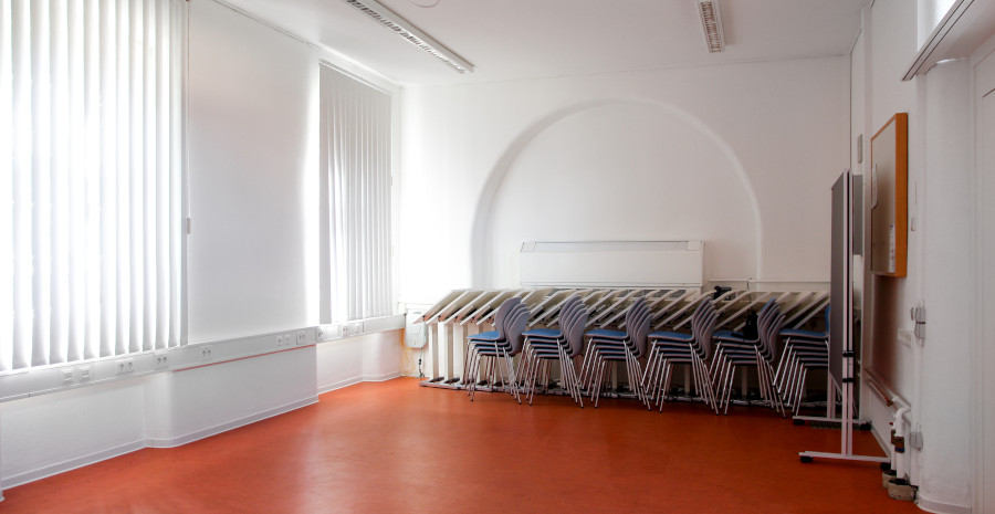 Foto von einem hellen, leeren Raum mit farbigem Boden an dessen Stirnseite Tagungsmöbel in zusammengeklappter und gestapelter Form bereitstehen.