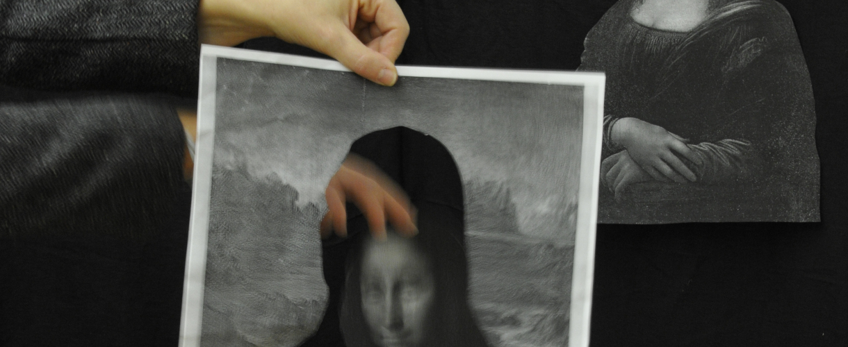 Foto: Zwei Hände halten eine Schwarz-Weiß-Kopie der Mona Lisa mit ausgeschnittenem Gesicht vor einem schwarzen Hintergrund, daneben hängt ein weiteres Bild der Mona Lisa – die Szene spielt spielerisch mit Identität, Aneignung und dem berühmten Kunstwerk.