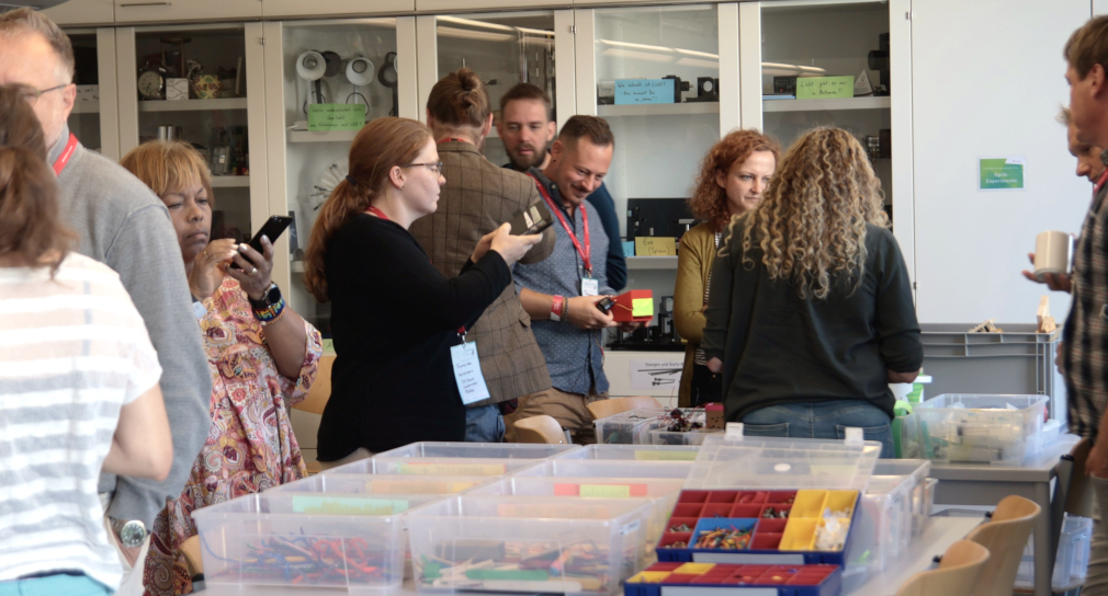 Foto: Eine Gruppe von Lehrkräften erkundet in einem Schulungsraum für Naturwissenschaften verschiedene Materialien in niedrigen Boxen, die auf Tischen stehen. Die insgesamt elf Lehrkräfte unterhalten sich oder machen mit ihren Smartphones Fotos von den Materialien.