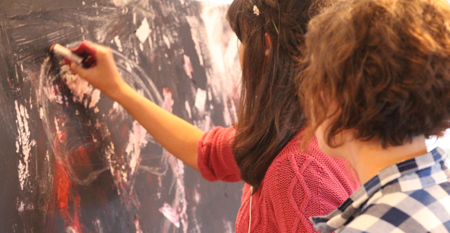 Link zu den Seiten der Stiftung Kinderland Baden-Württemberg; Foto: Zwei Mädchen wird beim Malen eines großen Bildes über die Schulter geschaut.